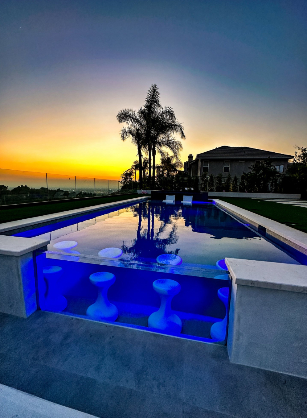 Illuminated acrylic pool wall with underwater bar stools at sunset, framed by palm trees